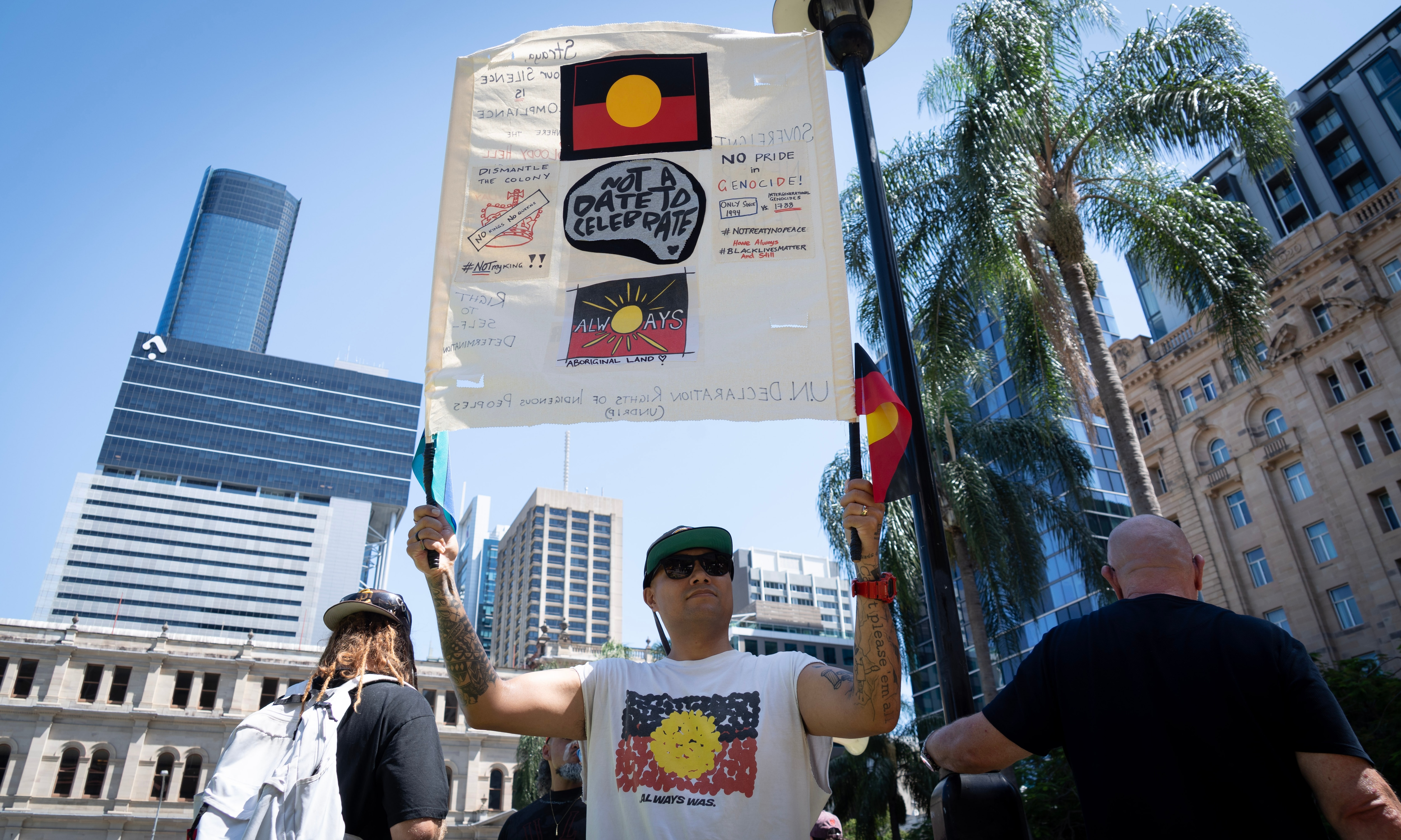 A man wearing a shirt featuring the Aboriginal flag holds up a sign.