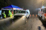 Police work in the rain near the Targa crash. A police car has its headlights on, shining through the rain.