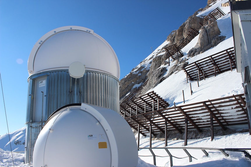 A tall telescope dome and a shorter one, each a couple of metres wide, with avalanche barriers on the slope behind