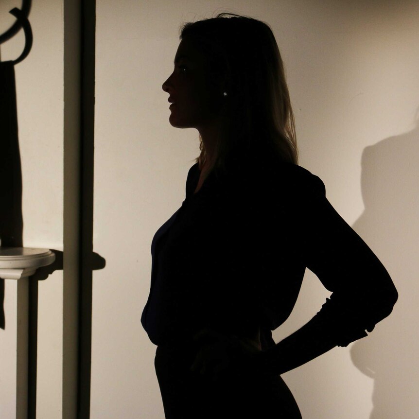 A woman stands silhouetted in shadow inside a bedroom.