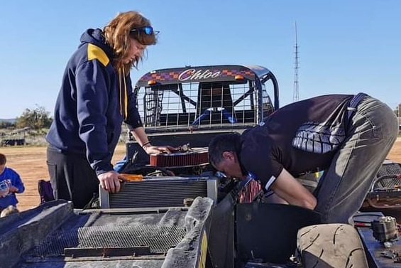 Man on right bent over engine of racecar fixing something, woman watching from left of car