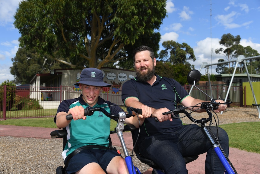 A young boy in unform and an adult ride on adapted tricycles outside a school playground