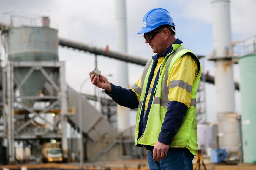 A man with a hardhat and reflector clothing on holding a small square, grey piece of concrete at a power station