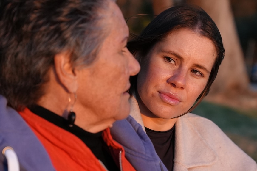 Two women sit and talk to each other. The younger woman is looking at the older woman’s face.  