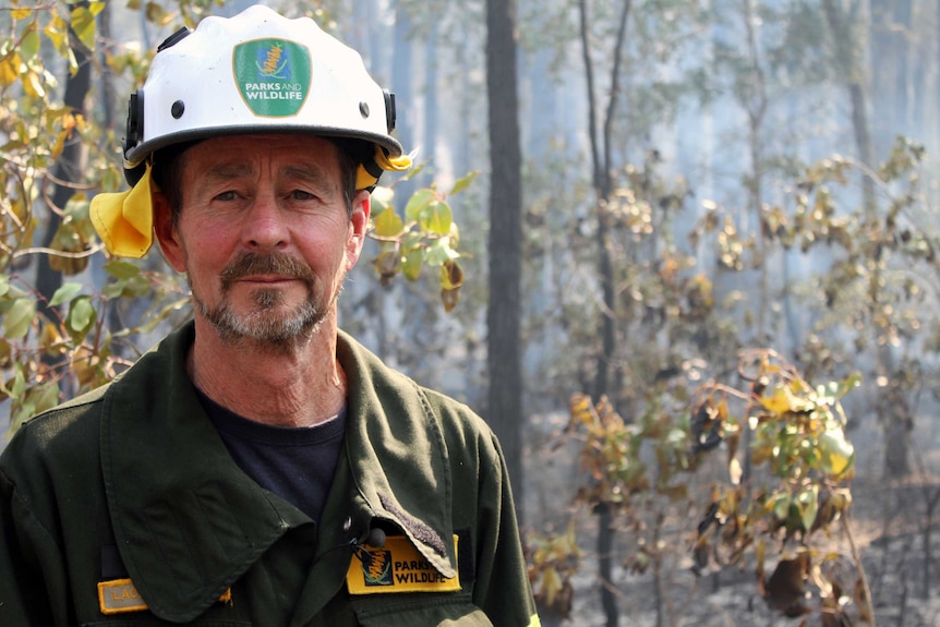 A man in a parks and wildlife jackets and fire helmet stands in burnt out bushland.