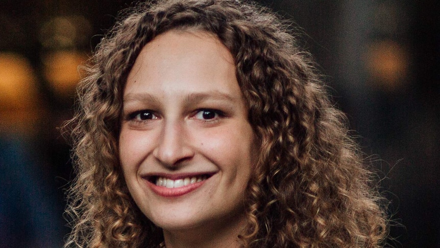 Moya Sarner, wearing white shirt and with curly brown shoulder-length hair, smiles widely.