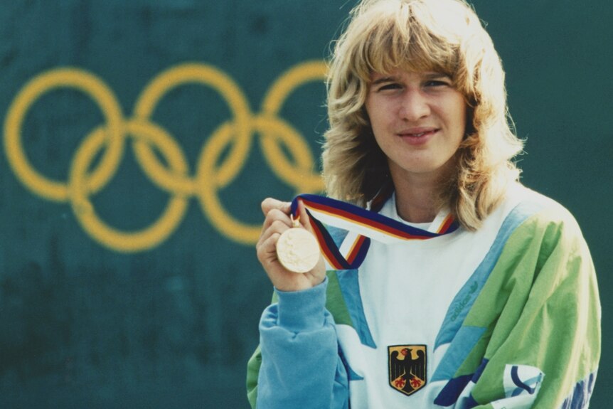 Steffi Graf holds up an Olympic gold medal in front of the Olympic rings.