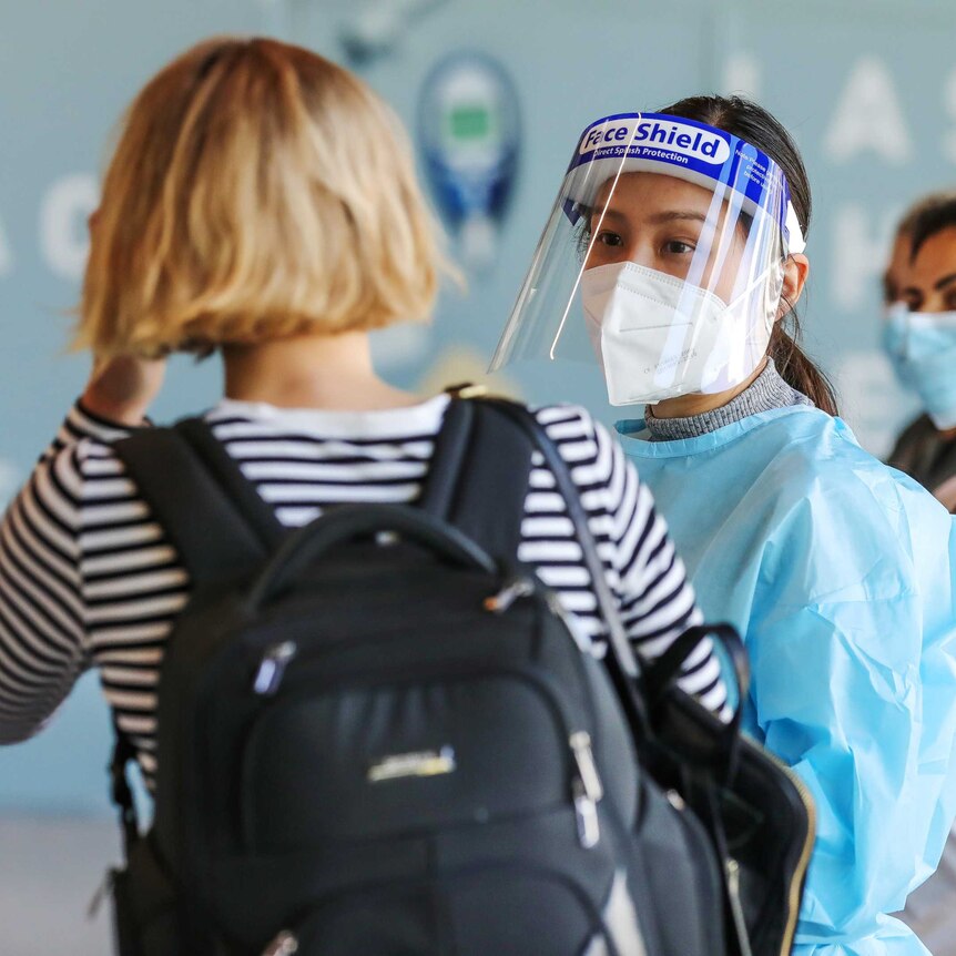 A medical worker in PPE wearing a mask and visor talks to a disembarked passenger at an airport