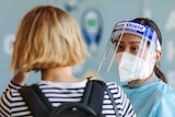 A medical worker in PPE wearing a mask and visor talks to a disembarked passenger at an airport
