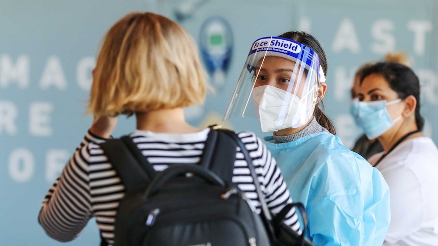 A medical worker in PPE wearing a mask and visor talks to a disembarked passenger at an airport