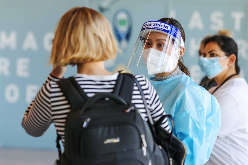 A medical worker in PPE wearing a mask and visor talks to a disembarked passenger at an airport