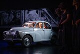 A blue car on a stage with actors inside