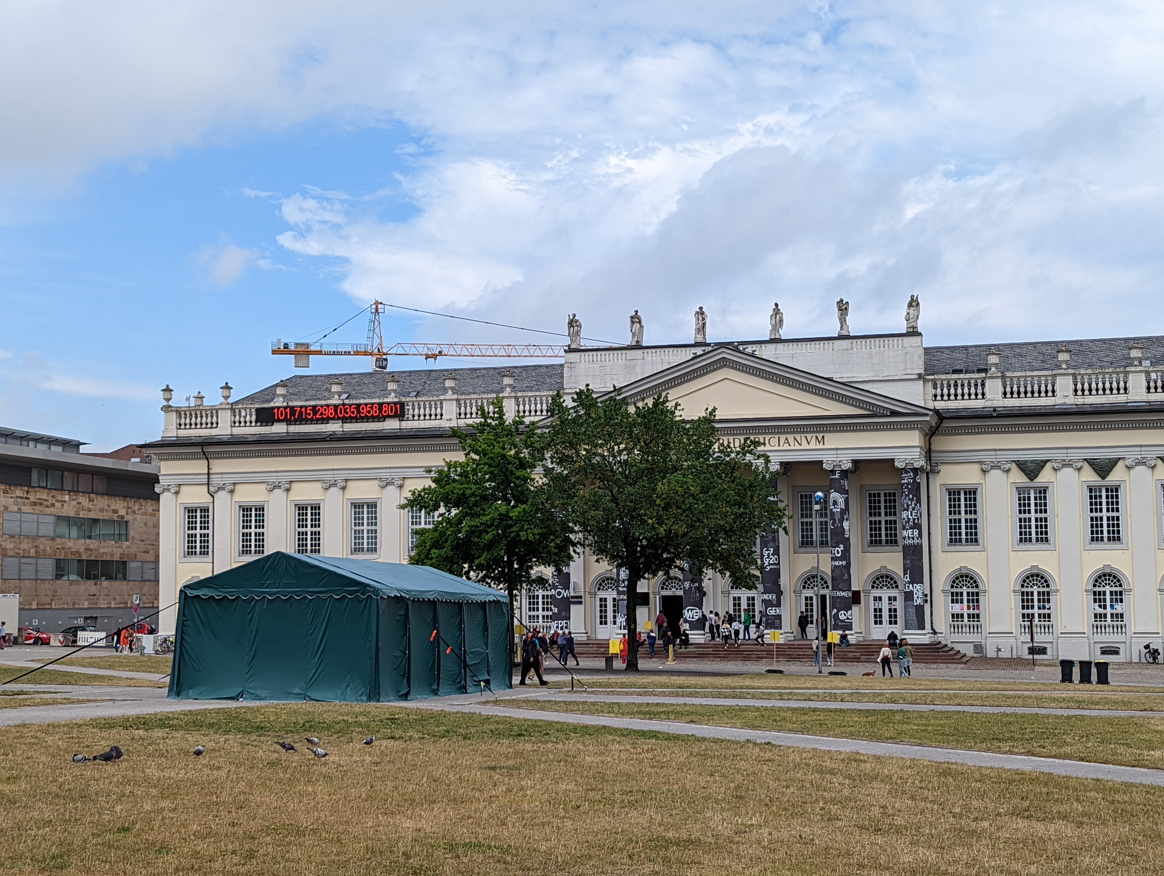 A rear view of a large green tent set in a grassy park in front of a large white German classicist style art gallery.