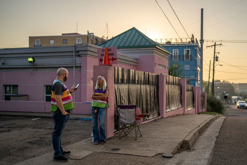 A man and a woman in brightly coloured vests stand outside a pink building talking at sunset 