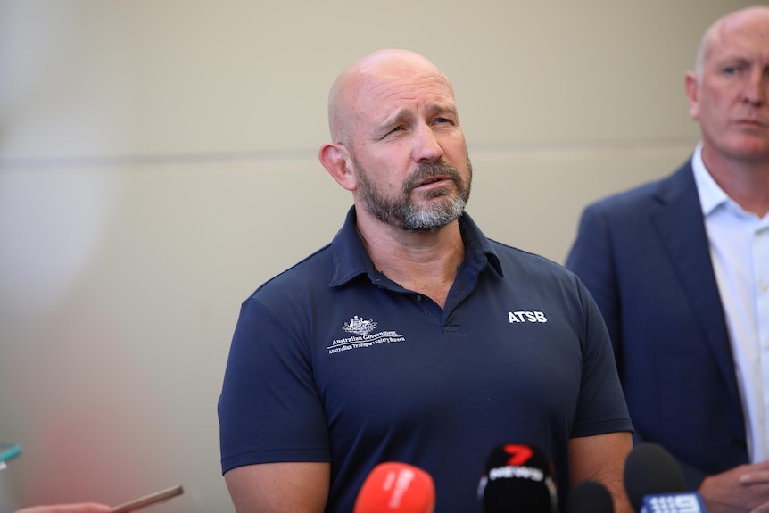 A man in a navy blue polo shirt speaking at a press conference. His shirt says 'ATSB' on it. 