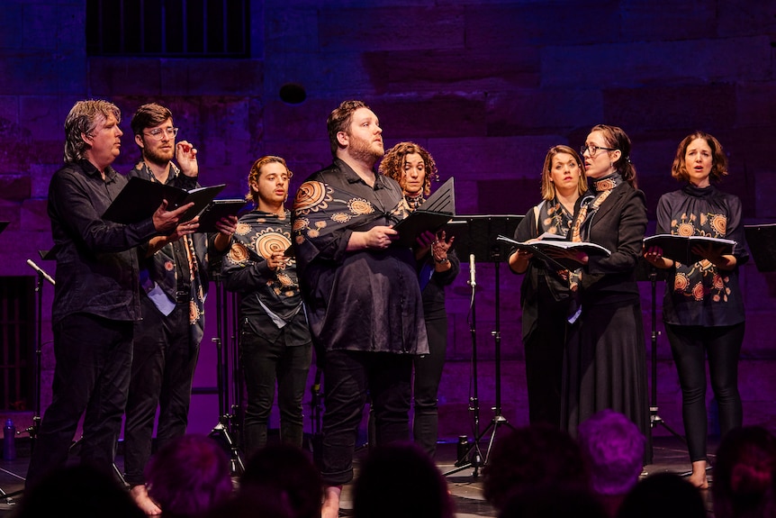 Eight singers in black except for colourful Indigenous scarves draped around them, gather together singing at front of stage. 