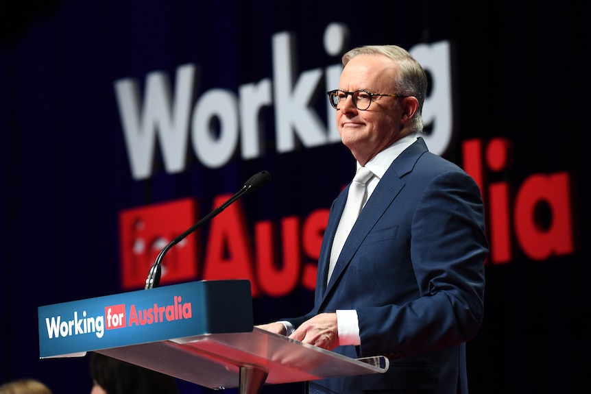 A male politican in a blue suit and white tie, speaking at a lecturn in front of a sign that reads 'working for Australia'