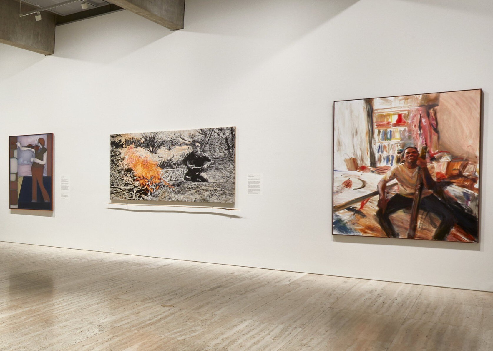 On a white gallery wall hangs three paintings, including one of an Indigenous man with a didgeridoo.
