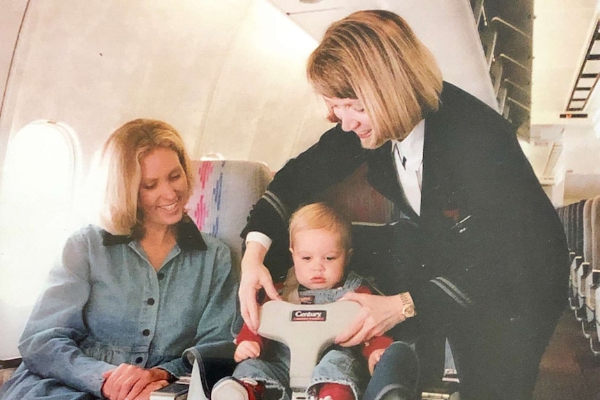 A flight attendant places a restraint over an infant in a car seat as a mother watches on in the cabin of an airplane.