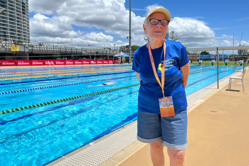 An elderly woman with black glasses in a blue t-shirt with an orange lanyard and denim shorts standing in front of a pool  