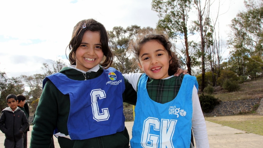 Two young girls wearing netball bibs over their school uniforms pose smiling with arms around one another on the netball court.