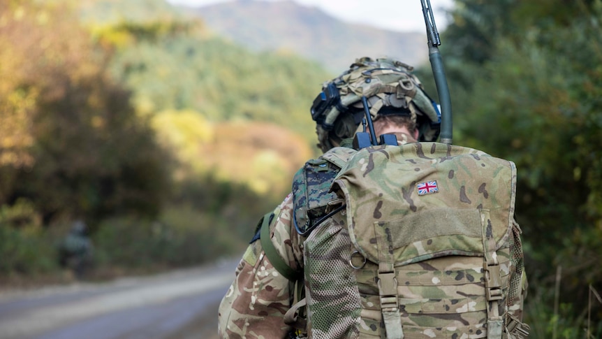 A soldier in camouflage and a backpack with the Union Jack has their back to the camera, green hills in the background