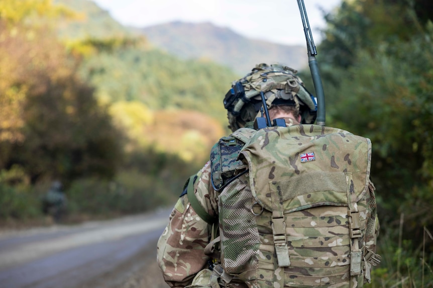 A soldier in camouflage and a backpack with the Union Jack has their back to the camera, green hills in the background
