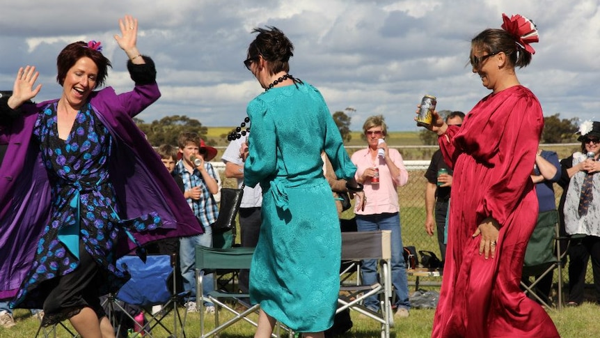 Three women are dancing, wearing colourful dresses. There are people standing and smiling in the background.