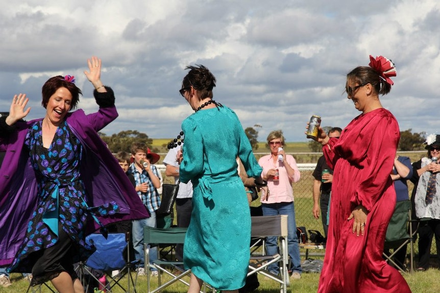 Three women are dancing, wearing colourful dresses. There are people standing and smiling in the background.