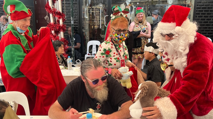 a man dressed as santa gives another man sitting down eating a christmas present