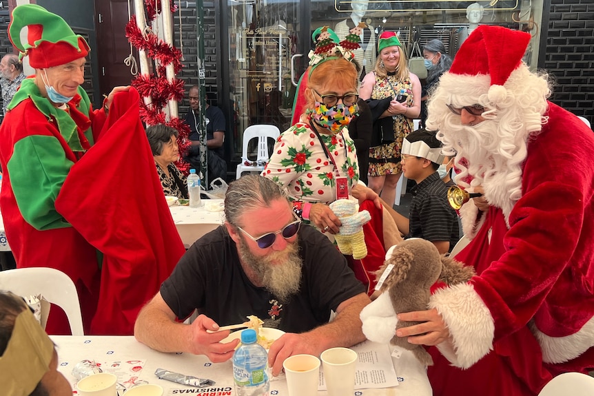 a man dressed as santa gives another man sitting down eating a christmas present