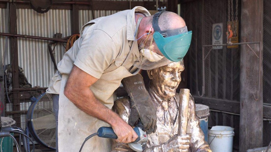 A man wearing a protective mask over his mouth and shield over his eyes buffs a bronze statue of a man with a power tool.