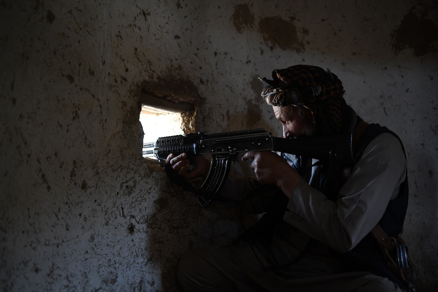 A man aims a rifle out the window of a small stone hut. 