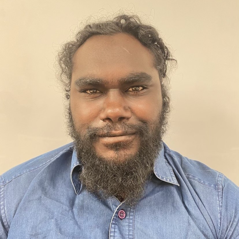 A man stares into the camera with a slight smile and a wall behind him.