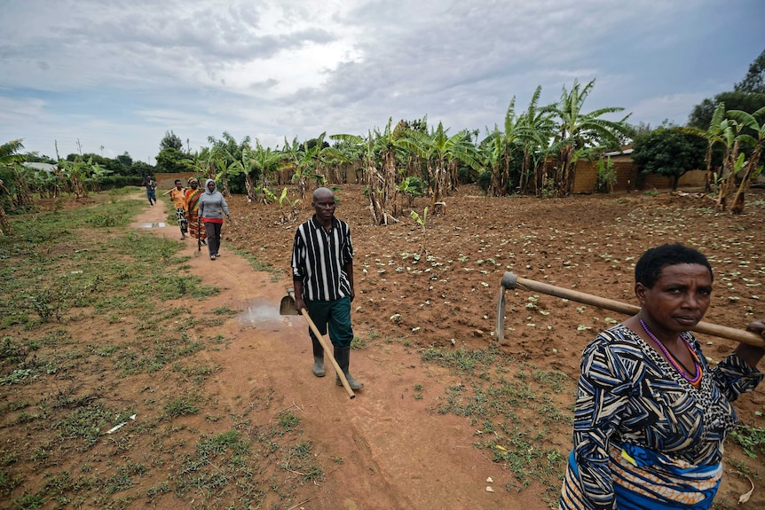 Frederick Kazigwemu walks carrying a hoe with others also walking in a field.