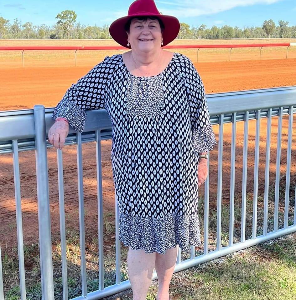 Una mujer mayor sonriente con un sombrero se encuentra frente a una pista de carreras en el campo.