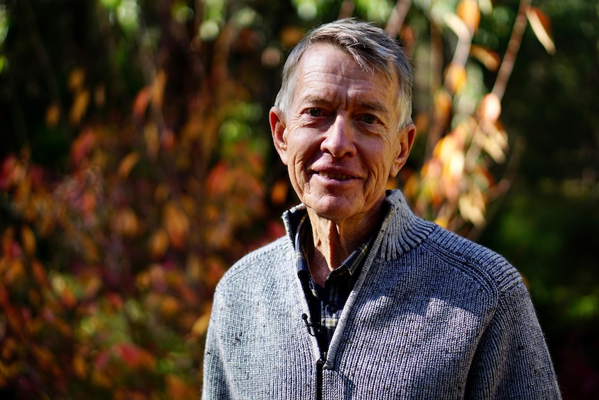 An older man with short grey hair, wearing a wool cardigan, stands in front of a leafy garden.