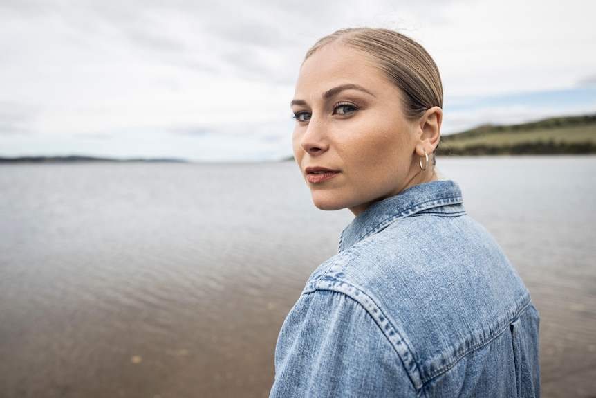 A woman in a denim shirt looks over her shoulder while standing in front of a river