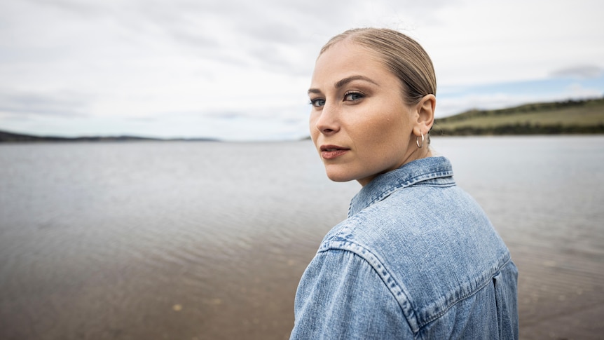 A woman in a denim shirt looks over her shoulder while standing in front of a river