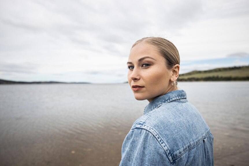 A woman in a denim shirt looks over her shoulder while standing in front of a river