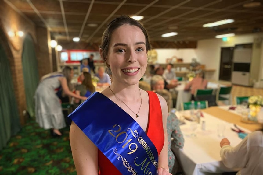 Girl in red dress in dining room wearing a blue ribbon sash.