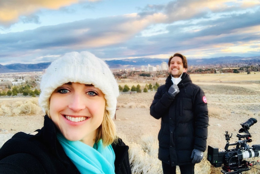 Cameraman and camera and Diss standing in desert with city skyline in background.