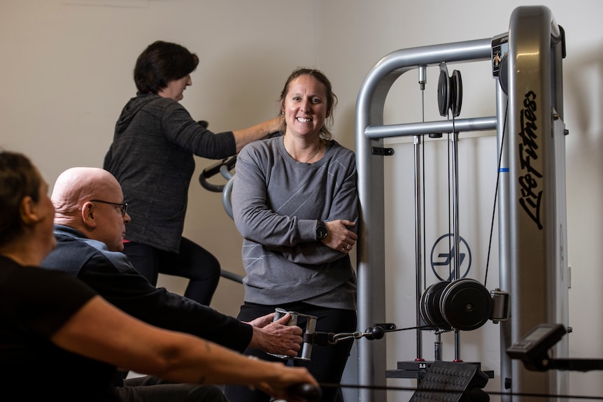 Nadia stands with her arms folded, leaning against a training machine at the gym. People are working out around her. 