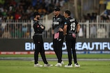 Three cricket players in black have a discussion in the middle of a cricket field in a night game