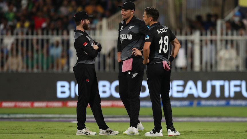 Three cricket players in black have a discussion in the middle of a cricket field in a night game