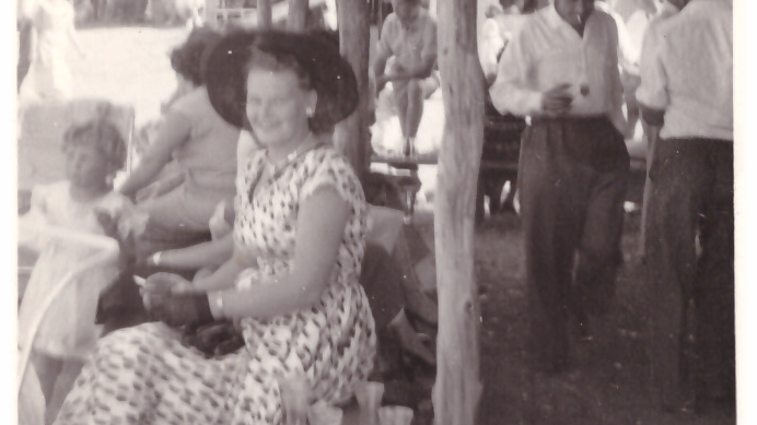 A blsack and white, grainy pic of a smartly-dressed lady in a patterned dress sitting on a bench, turning to smile at the camera