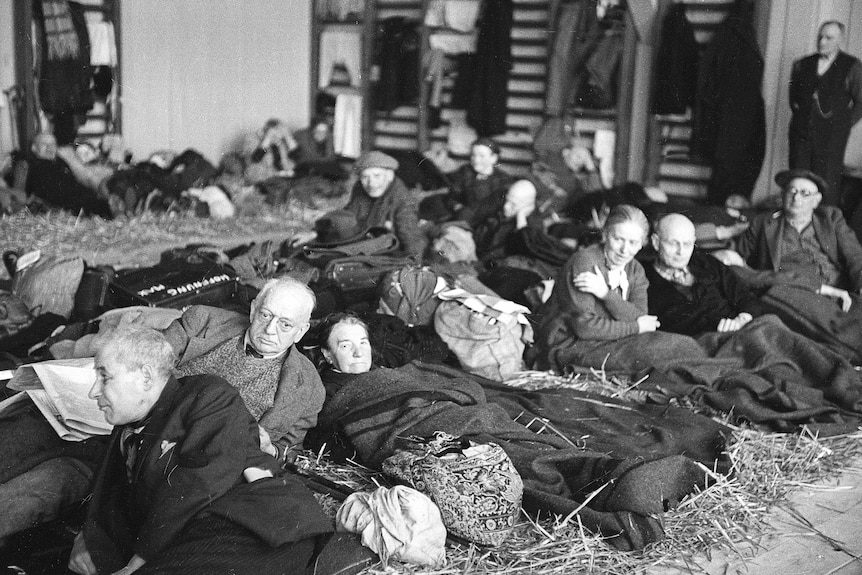 Black and white photo of a crowd of mainly older men and women laying on the floor wrapped in blankets with serious expressions.