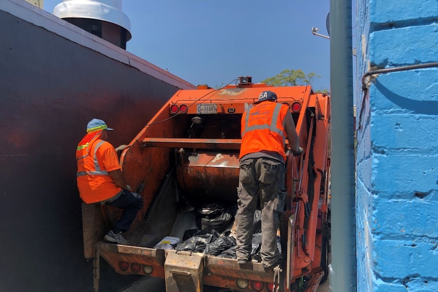 Two men are seen on the back of a rubbish truck in a narrow lane.