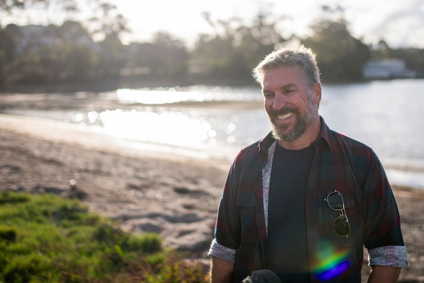 A man in a plaid shirt stands on the shore of a beach smiles with the ocean and bay in the background.