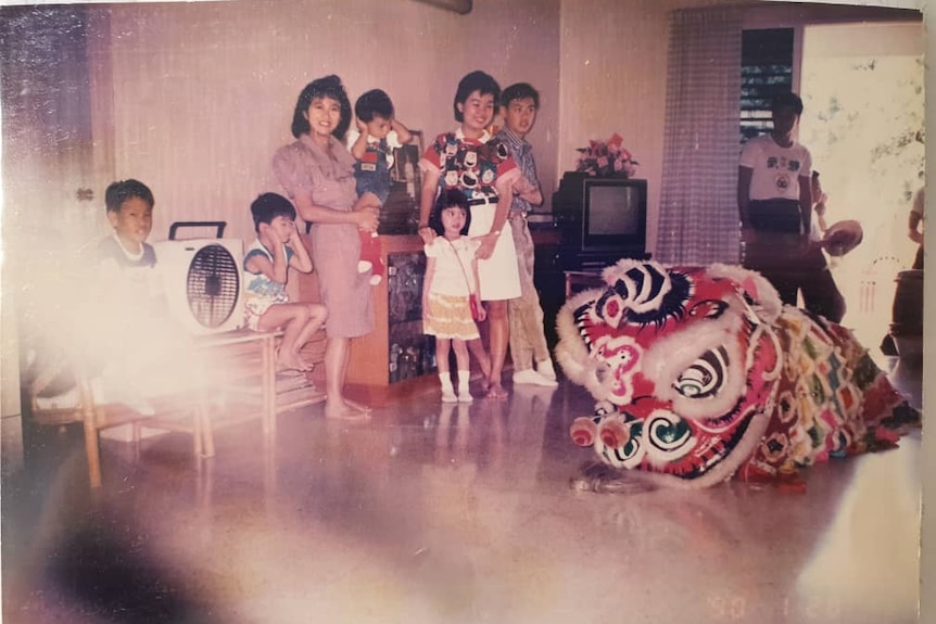 Four children are seen standing amongst four adults in a 90s lounge room against a wall, with a dragon costume on the ground